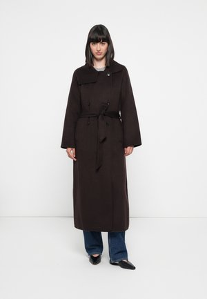 Brown long coat with a wrap design, wide sleeves, two buttons, and a belt at the waist. Worn over blue jeans and black shoes.