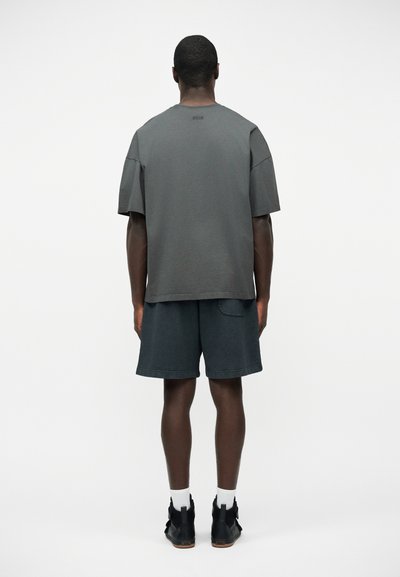 Homme portant un t-shirt gris foncé oversize, un short assorti, des chaussettes blanches et des chaussures noires, debout de dos sur un fond uni.