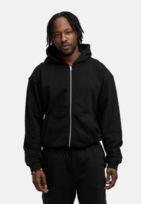 Schwarze Zip-Hoodie aus weichem Stoff, mit einer geräumigen Kapuze, Känguru-Taschen und gerippten Bündchen. Moderner, lässiger Schnitt.