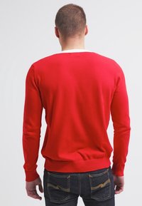 Roter Strickpullover mit langen Ärmeln, geribbelten Manschetten und Saum, rundem Ausschnitt und einem weißen Akzent am Kragen, getragen über dunklen Jeans.
