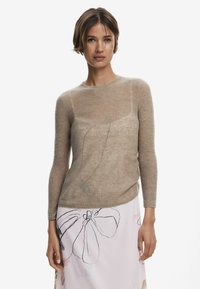 Beige, ribbeliger Strickpullover mit langen Ärmeln und körperbetontem Design, über einem hellen Rock mit floralem Linienmuster getragen.