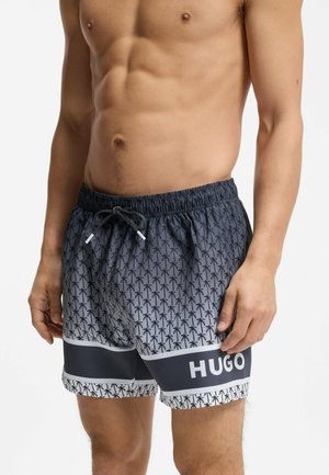 Shirtless man wearing gray swim shorts with black palm tree pattern and black stripe featuring white "HUGO" logo, standing with relaxed arms.