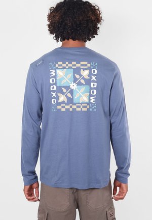 Personne aux cheveux bouclés portant une chemise à manches longues bleue avec un motif floral et à carreaux en forme de carré, avec le texte "OXBOW" dans le dos.