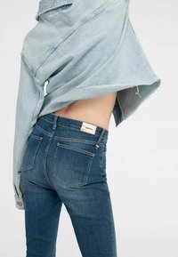 Giacca di jeans blu chiaro appoggiata su jeans blu scuro con cinque tasche, caratterizzata da una texture sbiadita e dettagli in metallo argentato.