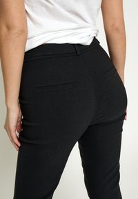 Person wearing black fitted pants and white shirt shown from the waist down, focusing on the back and hand resting by the side.