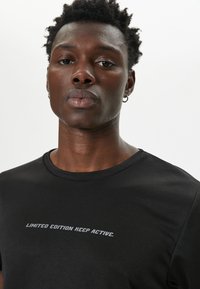 Camiseta negra con cuello redondo, que presenta una impresión de texto horizontal "EDICIÓN LIMITADA MANTENTE ACTIVO" en gris a lo largo del pecho.