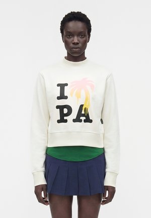 Άτομο με σκουρόχρωμο δέρμα που φοράει λευκό φούτερ με την επιγραφή "IPA" και μια παστέλ εικόνα φοίνικα, πράσινο πουκάμισο από κάτω και μια μπλε πλισέ φούστα.