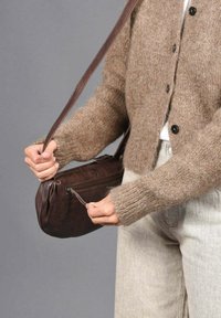 Bolso de mano de cuero marrón con una textura suave, que cuenta con un cierre de cremallera, una correa larga y una forma redondeada, sostenido por una persona que lleva un suéter claro.