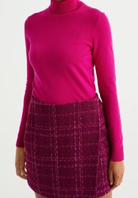 Maglione a collo alto fucsia in tessuto soffice abbinato a una gonna rosa scuro testurizzata con un leggero motivo a quadretti e cuciture visibili.