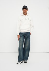 Sudadera blanca con logo en relieve, combinada con jeans de pierna ancha azules que presentan detalles de patchwork y zapatillas plateadas con acentos reflectantes.