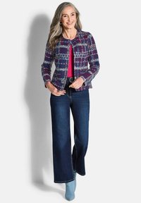 Strickjacke aus lila, blauer und grauer gemusterter Stoff, kombiniert mit einem roten Oberteil und dunkelblauen, ausgestellten Jeans. Blaue spitze Stiefel runden das Outfit ab.