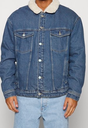 Homme portant une veste en denim bleu avec col en polaire blanche, fermeture par boutons, poches poitrine à rabat, et jean bleu clair, debout les mains détendues.