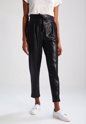 Frau trägt schwarze, glänzende High-Waist-Hose mit Kordelzug und weißen Sneakers, steht vor einem schlichten hellen Hintergrund.