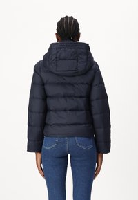 Persona con capelli intrecciati che indossa una giacca imbottita nera con cappuccio e jeans blu, vista di spalle su sfondo bianco.