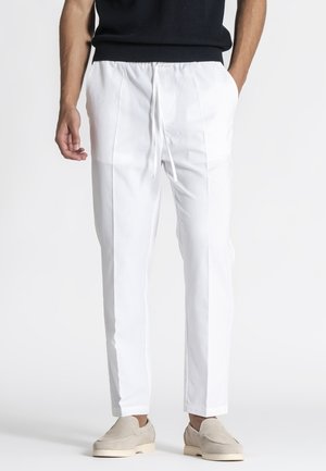 Muž v bielych na mieru ušitých nohaviciach so sťahovacou šnúrkou, béžových slip-on topánkach a čiernom tielku bez rukávov, stojaci s rukami čiastočne v vreckách.