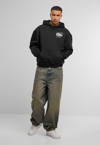 Felpa nera con grande tasca frontale e stampa grafica. Abbinata a jeans larghi e sbiaditi di colore verde e sneakers bianche. Design semplice e informale.