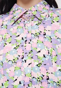 Blusa floral multicolorida com colarinho, apresentando padrões em rosa, roxo e verde sobre um fundo claro. Inclui detalhes em hardware dourado.