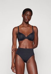 Vrouw met kort haar die een zwarte onderbuste beha en hoge zwarte onderbroek draagt, staand tegen een effen lichte achtergrond.