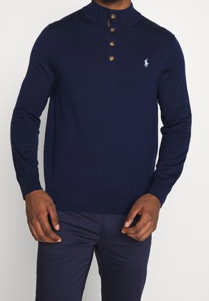 Uomo che indossa un pullover a maniche lunghe blu navy con tre bottoni marroni al collo e logo ricamato azzurro chiaro sul petto.