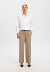 Camicia bianca con bottoni e una tasca singola, abbinata a pantaloni beige su misura. Tacchi neri a punta completano l'outfit.