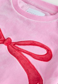 Różowy pluszowy sweatshirt z czerwonym aksamitnym wzorem kokardy na przodzie. Okrągły dekolt i miękka faktura na całej powierzchni.