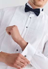 Biała koszula z plisowanym przodem, czarnymi guzikami i francuskimi mankietami. Wykończona czarnym krawatem w muszkę i teksturowanym materiałem.
