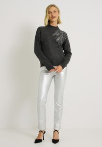 Pull gris à col montant, avec un motif de strass sur un côté, associé à un pantalon skinny argenté brillant et des talons noirs.