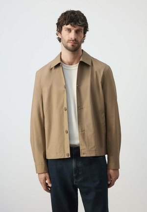 Mann mit kurzen lockigen Haaren, der eine beige Hemdjacke über einem weißen Hemd und dunkelblauen Jeans trägt, steht vor einem einfarbigen hellen Hintergrund.