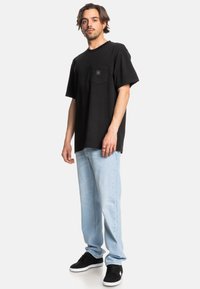Černé bavlněné tričko s kapsou na hrudi, v kombinaci s světle modrými džínami a černými teniskami s bílou podrážkou, postavené na bílém pozadí.