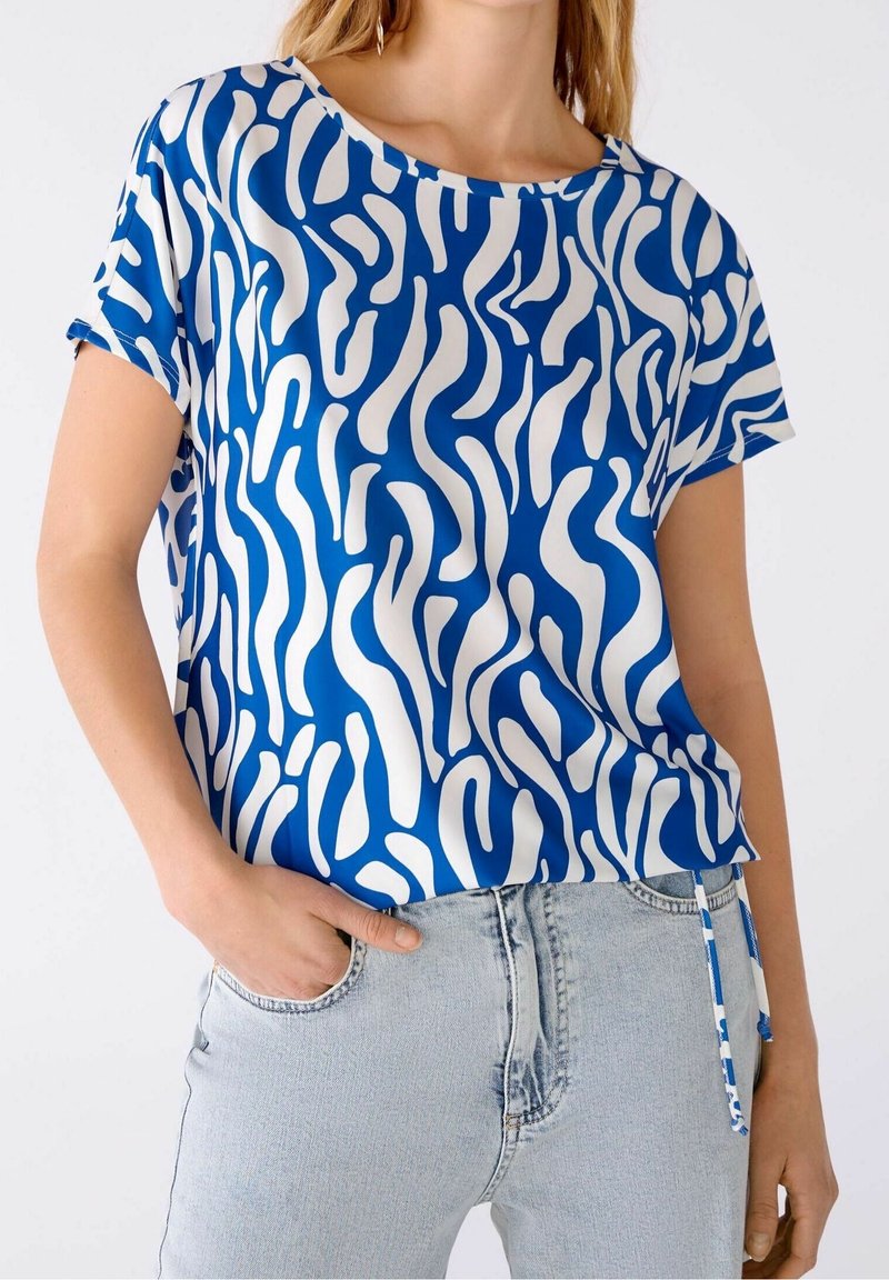 Blå og hvid bluse med et flydende, bølget mønster, korte ærmer og en rund halsudskæring, sat sammen med lyse denim jeans.
