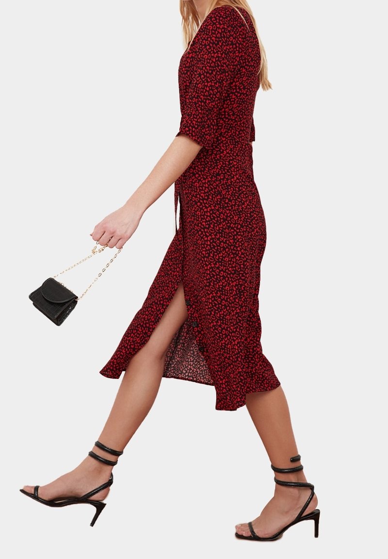 Femme marchant dans une robe rouge et noire à imprimé léopard avec une fente devant, tenant une petite pochette noire, portant des talons noirs à brides.