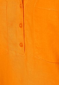 Een oranje linnen shirt met een knoopsluiting aan de voorkant, twee borstzakken en zichtbare stiksels. Soepele textuur en levendige kleur.