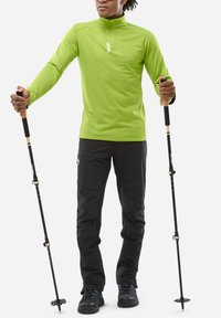 Haut de sport vert à manches longues avec une fermeture éclair quart, en tissu texturé. Pantalons noirs et bâtons de randonnée avec poignées en bois, chaussures robustes.