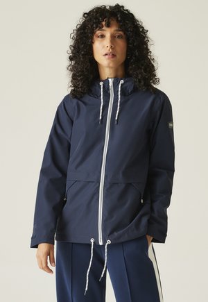 Femme aux cheveux bouclés portant une veste à capuche bleu marine avec une fermeture éclair blanche et des cordons, assortie à un pantalon bleu avec des rayures blanches.