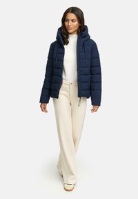 Marine blauwe pufferjack met kap, gequilt patroon en koord, gedragen over een witte top en crèmekleurige wijde broek. Casual schoenen maken de look compleet.