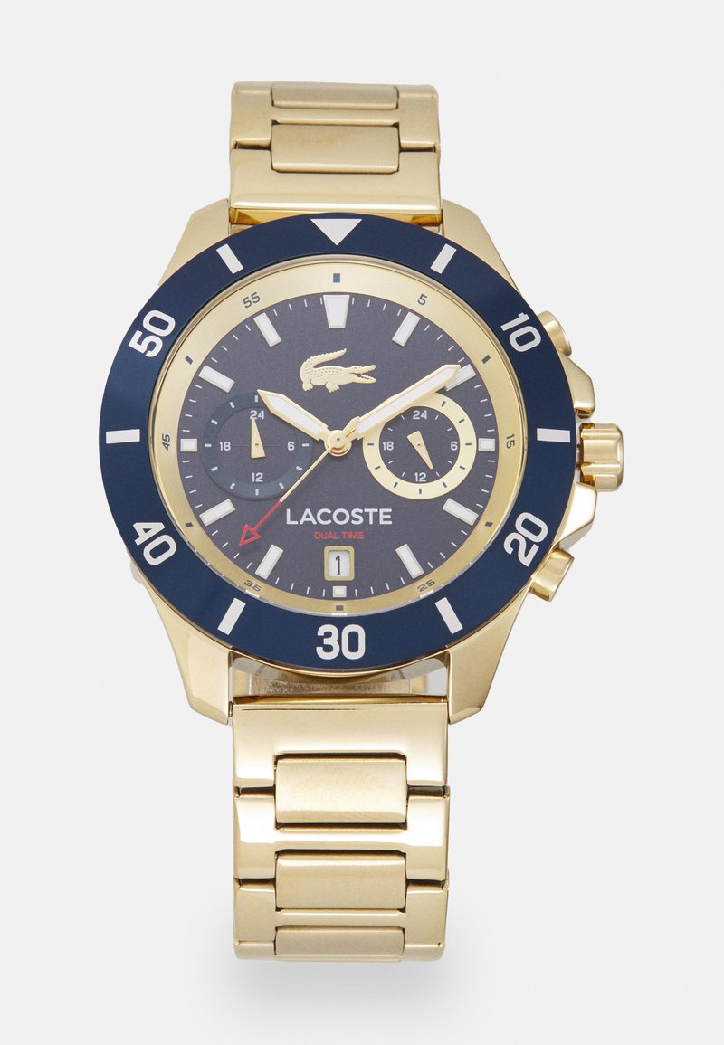 Reloj de oro con un bisel azul marino, una esfera azul marino y marcadores de horas blancos. Presenta subesferas y una pulsera de eslabones metálicos. Logotipo de Lacoste mostrado.