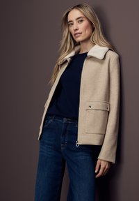 Blondýnka v béžovém saku s bílým límcem, pod kterým má námořnický svetr, a tmavě modrých džínách, opírající se o mauve zeď.