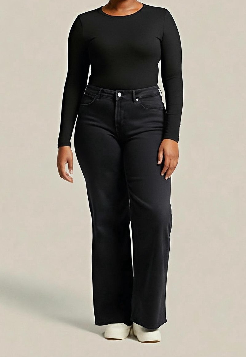 Personne portant un haut noir moulant à manches longues, un jean noir taille haute à jambes larges et des chaussures blanches, debout devant un fond beige.