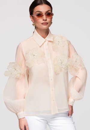 Femme portant un chemisier rose clair transparent avec de grandes applications florales sur les manches et la poitrine, associé à un pantalon blanc et des lunettes de soleil teintées marron.