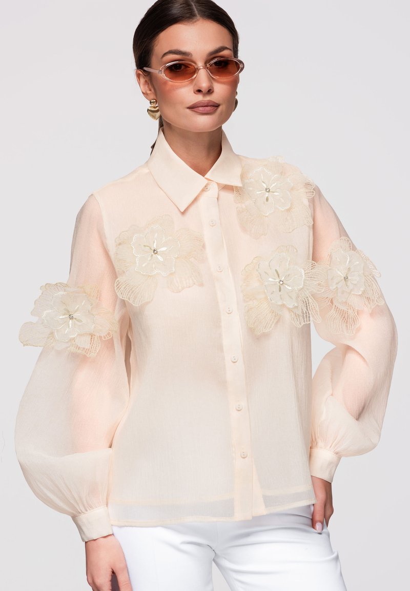 Femme portant un chemisier rose clair transparent avec de grandes applications florales sur les manches et la poitrine, associé à un pantalon blanc et des lunettes de soleil teintées marron.