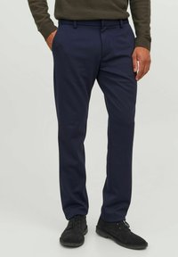 Pantalones de traje azul marino en una tela suave con corte recto, que incluyen trabillas para el cinturón y bolsillos laterales, combinados con zapatos de ante negros.