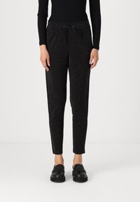 Pantalon noir fuselé en tissu pailleté, avec une taille élastique dotée d'un cordon de serrage et des poches latérales. Porté avec des chaussures noires à enfiler.