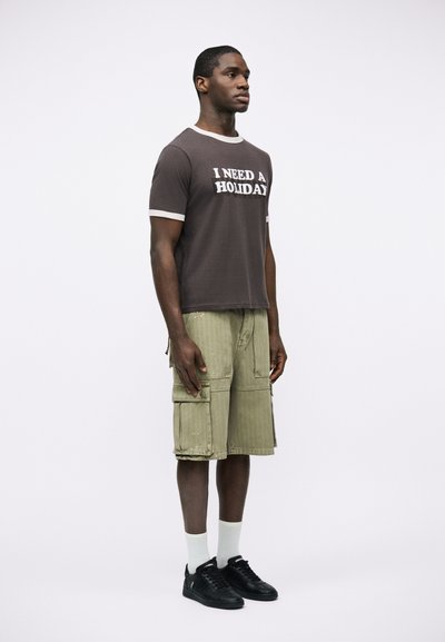 Νέος άνδρας που στέκεται, φοράει σκούρο μπλουζάκι με το κείμενο "ΧΡΕΙΑΖΟΜΑΙ ΔΙΑΚΟΠΕΣ", πράσινα cargo σορτς, λευκές κάλτσες και μαύρα αθλητικά παπούτσια.
