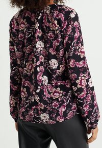 Žena v černé blůze s dlouhým rukávem a růžovým a bílým květinovým vzorem, v kombinaci s černými koženými kalhotami, zobrazená zezadu.