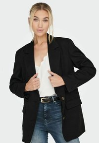 Blazer nero in tessuto texturizzato, caratterizzato da una vestibilità su misura, grandi tasche frontali e bottoni dorati. Indossato sopra una maglietta bianca e jeans.