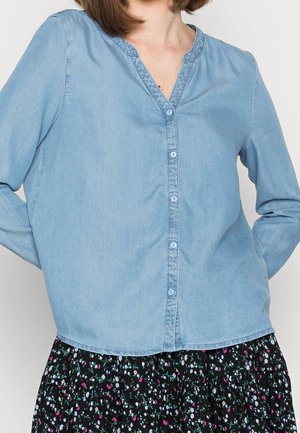 Lichtblauw denim overhemd met knopen, een V-hals, lange mouwen en een gebogen zoom. Gecombineerd met een zwarte rok met bloemenpatroon.