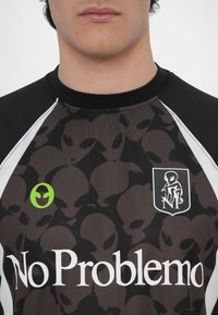 Camiseta de manga curta preta e castanha com padrões de alienígenas, acabamentos brancos, logótipo verde de alienígena e texto "No Problemo" em fonte branca em negrito.