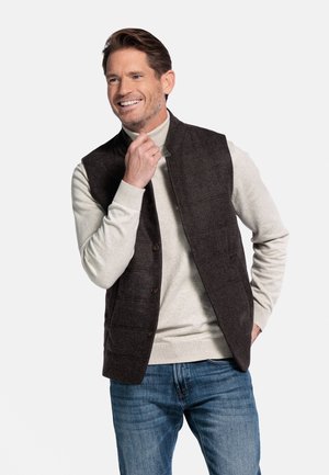 Dark brown quilted vest with large buttons, worn over a light grey turtleneck. Model poses casually in blue jeans against a plain background.