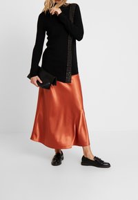 Czarny sweter z długim rękawem z metalowymi detalami po bokach, zestawiony z błyszcząca pomarańczową satynową spódnicą midi i czarnymi butami na slip-on, trzymająca czarną kopertówkę.