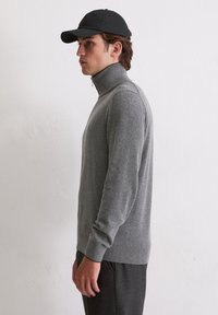 Grauer Rollkragensweater mit langen Ärmeln, aus weichem Stoff, kombiniert mit einer schwarzen Mütze, getragen mit dunklen Hosen. Seitenansicht.